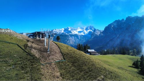 Green alpine slope with ski lift poles and a small alpine hut, snow-covered mountains and clear blue sky in the background.
