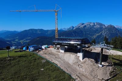 Aerial view of a ski lift station under construction on a mountain, with a crane, construction machinery and distant mountains in the background.