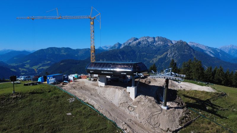 Luftaufnahme einer im Bau befindlichen Skiliftstation auf einem Berg, mit einem Kran, Baumaschinen und entfernten Bergen im Hintergrund.