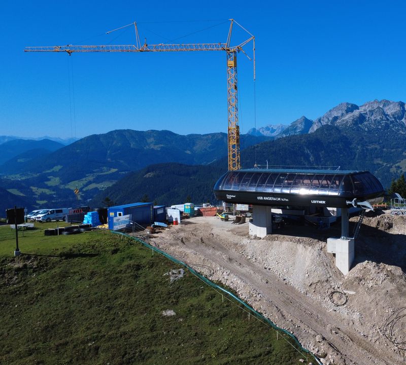 Aerial view of a ski lift station under construction on a mountain, with a crane, construction equipment, and distant mountains in the background.