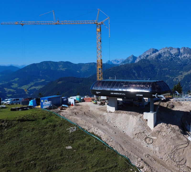 Luftaufnahme einer im Bau befindlichen Skiliftstation auf einem Berg, mit einem Kran, Baumaschinen und entfernten Bergen im Hintergrund.