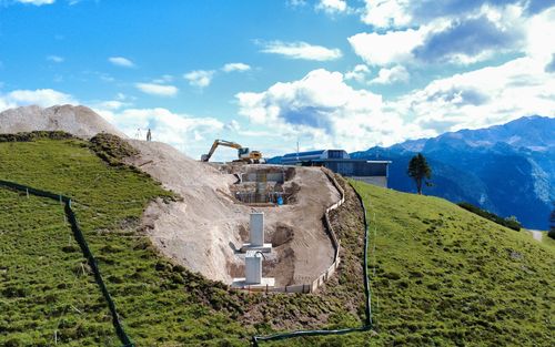 Construction site on a grassy hill with an excavator, piles of earth and concrete structures. Mountains and cloudy sky in the background.