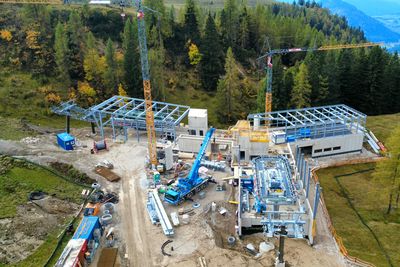 Luftaufnahme einer Baustelle in einer Bergregion mit Kränen, Stahlkonstruktionen und umgebenden Bäumen in Herbstfarben.