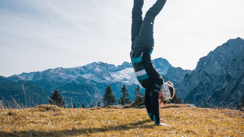 Person macht Handstand auf goldener Wiese vor schneebedeckten Bergen unter hellem Himmel.