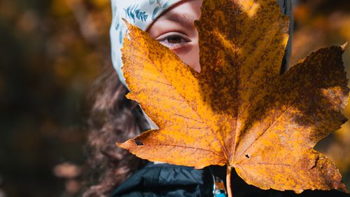 Ein Kind mit Stirnband hält ein großes, gelb-braunes Herbstblatt vor das Gesicht, nur ein Auge ist sichtbar. Herbstlicher Hintergrund.