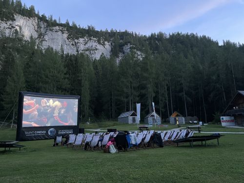 Evening open-air cinema on a meadow, featuring a screen and deckchairs set against a backdrop of wooded mountains.