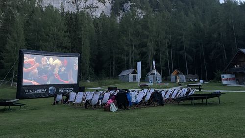 Abendliches Freilichtkino auf Wiese mit Leinwand und Liegestühlen vor bewaldeter Bergkulisse.