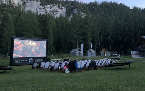 Abendliches Freilichtkino auf Wiese mit Leinwand und Liegestühlen vor bewaldeter Bergkulisse.