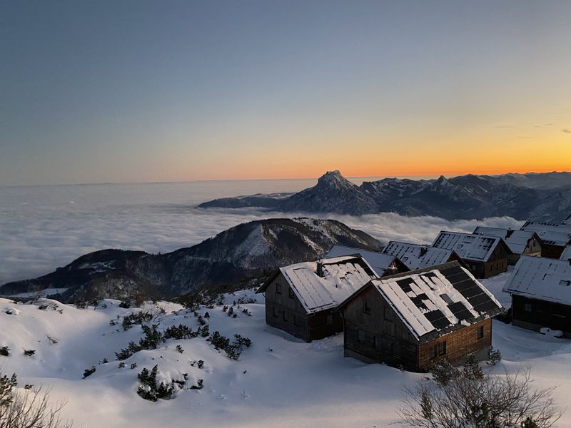 Snow-covered huts on the mountainside overlook a sea of clouds at sunset, with silhouetted peaks and a colorful sky in the background – Feuerkogel mountain village.