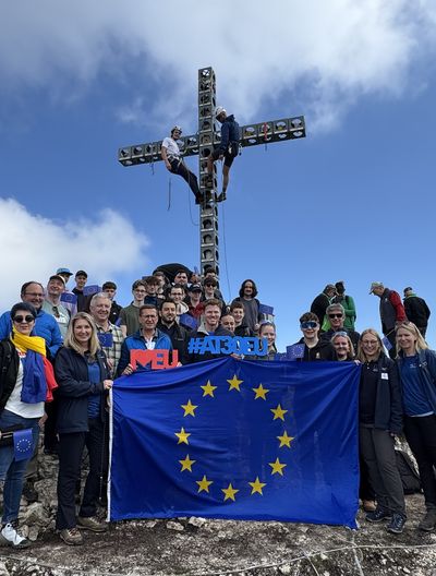 Junge Erwachsene mit EU-Flagge und #AT30EU-Schriftzug beim Gipfelkreuz am Feuerkogel unter blauem Himmel.