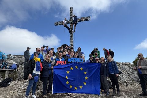 Junge Erwachsene mit EU-Flagge und #AT30EU-Schriftzug beim Gipfelkreuz am Feuerkogel unter blauem Himmel.