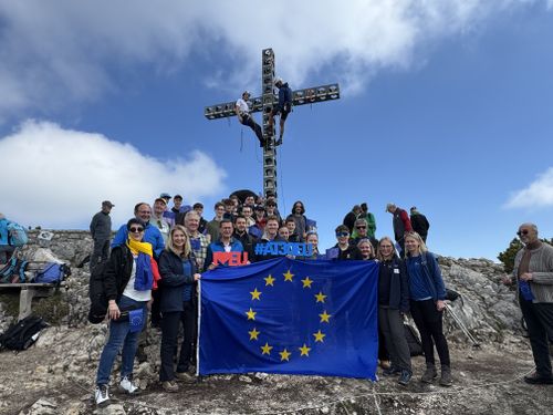 Junge Erwachsene mit EU-Flagge und #AT30EU-Schriftzug beim Gipfelkreuz am Feuerkogel unter blauem Himmel.