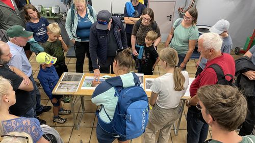 Eine Gruppe von Erwachsenen und Kindern steht um einen Tisch herum, während ein Mann auf Fotos zeigt und spricht.