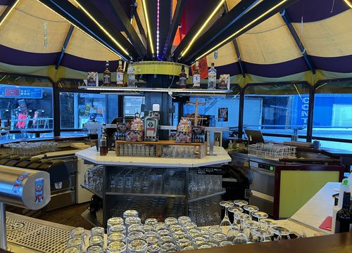 Runde Bar mit buntem Vordach, ordentlich angeordneten Gläsern und ausgestellten Flaschen. Große Fenster zeigen den Blick nach draußen auf die Straße.