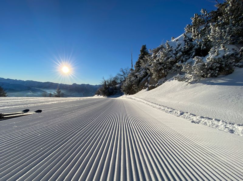 Tiefstehende Sonne über frisch präparierter Skipiste, daneben verschneite Bäume und Blick ins winterliche Tal.