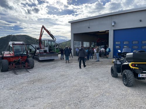 Eine Gruppe von Menschen steht auf einem Schotterplatz vor einem Gebäude, umgeben von einem roten Traktor, einem Bagger und einem ATV. Berge im Hintergrund.