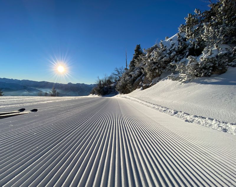 Strahlende Morgensonne über perfekt präparierter Skipiste mit Blick auf verschneite Berglandschaft und einzelne Skispitzen am Rand
