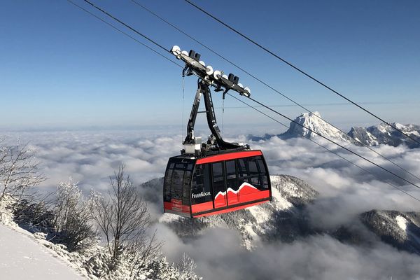 Eine rote Feuerkogel-Gondel schwebt vor verschneiten Gipfeln und einem Wolkenmeer mit Blick auf den markanten Traunstein.