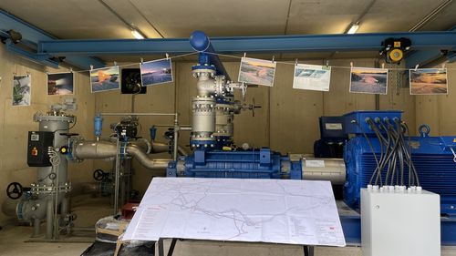 Industrielle Pumpstation mit blauen Wasserpumpen und grauen Rohren. Eine Karte liegt auf einem Tisch, Fotos von Schneekanonen hängen.