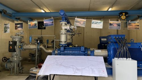 Industrielle Pumpstation mit blauen Wasserpumpen und grauen Rohren. Eine Karte liegt auf einem Tisch, Fotos von Schneekanonen hängen.