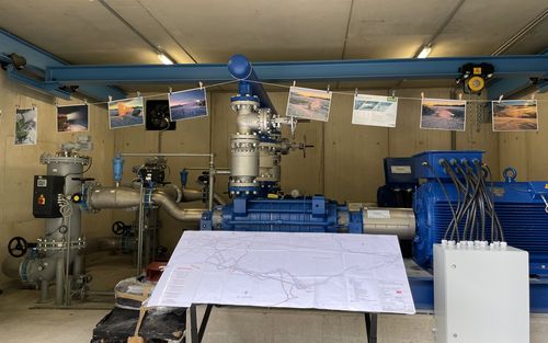 Industrielle Pumpstation mit blauen Wasserpumpen und grauen Rohren. Eine Karte liegt auf einem Tisch, Fotos von Schneekanonen hängen.