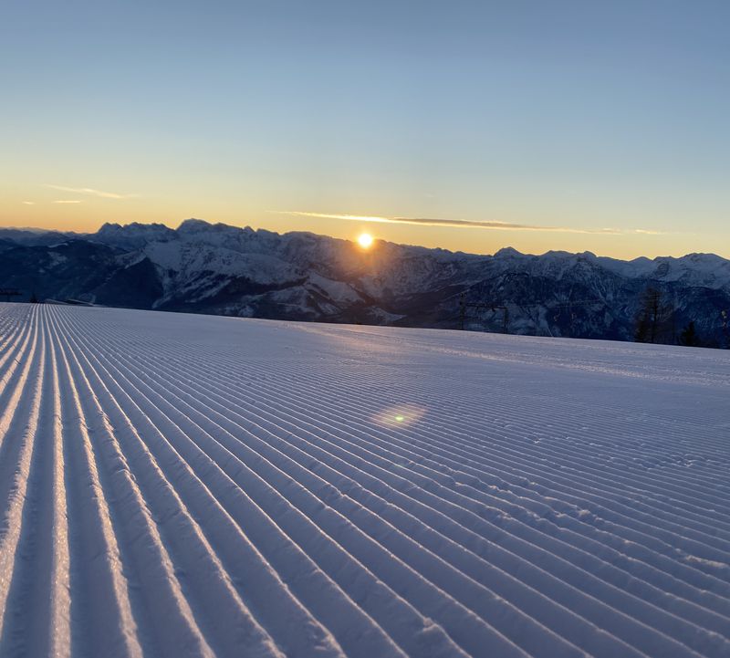 Die Sonne geht über verschneiten Bergen auf, im Vordergrund eine frisch gespurte Skipiste mit parallelen Linien unter klarem Himmel – Feuerkogel.