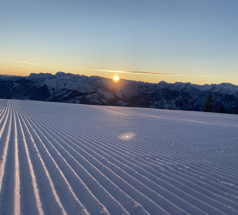 Erste Sonnenstrahlen tauchen die frisch präparierte Skipiste in warmes Licht, dahinter die stillen Berge.