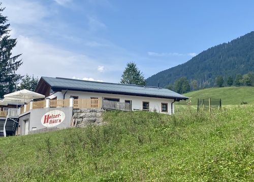 A small building with a sign reading ‘Holzer Hütte’, white parasols on a grassy hill, surrounded by trees and mountains under a clear blue sky.