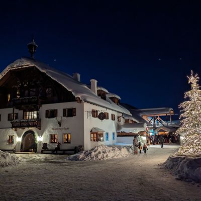 Winternacht mit schneebedecktem Landhaus, geschmückt mit Lichterketten, beleuchtetem Weihnachtsbaum und Menschen auf einem verschneiten Platz.