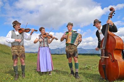 Vier Musiker in traditioneller bayerischer Kleidung spielen im Freien Instrumente, darunter Geigen, ein Akkordeon und einen Kontrabass, mit den Bergen im Hintergrund.