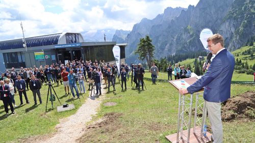 Ein Mann spricht auf einem Podium im Freien zu einer Menschenmenge, mit Bergen und einem Seilbahngebäude im Hintergrund unter einem teilweise bewölkten Himmel.