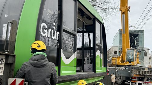 Drei Arbeiter mit gelben Schutzhelmen sichern eine große grün-schwarze Gondelkabine mit der Aufschrift „Grün“, neben der ein gelber Kran steht.