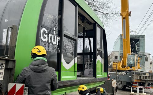 Drei Arbeiter mit gelben Schutzhelmen sichern eine große grün-schwarze Gondelkabine mit der Aufschrift „Grün“, neben der ein gelber Kran steht.