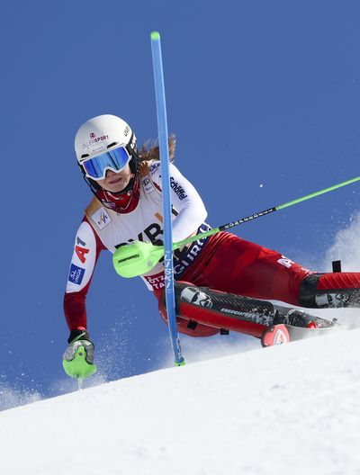 A female ski racer rounds a gate pole in a deep lean under a blue sky, kicking up a large spray of snow.