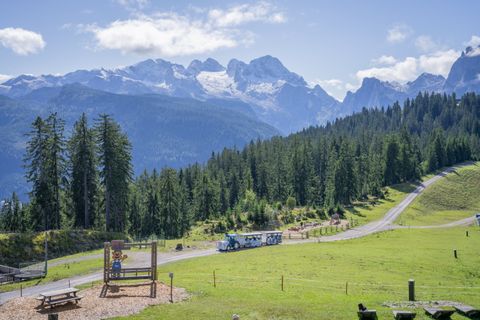 Ein malerischer Blick auf den schneebedeckten Dachstein Gletscher und einen Bummelzug, der auf einer kurvenreichen Straße fährt. Rundherum üppig grüne Bäume und Almwiesen unter blauem Himmel.
