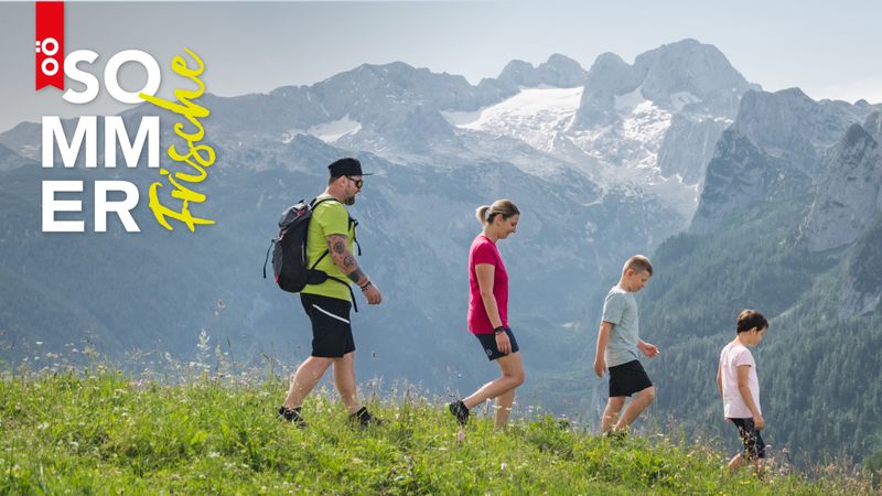 Familie beim Wandern auf einem grasbewachsenen Hügel mit dem Dachstein Gletscher im Hintergrund unter einem klaren Himmel. Text bzw. Logo auf dem Bild: „OÖ Sommerfrische.“