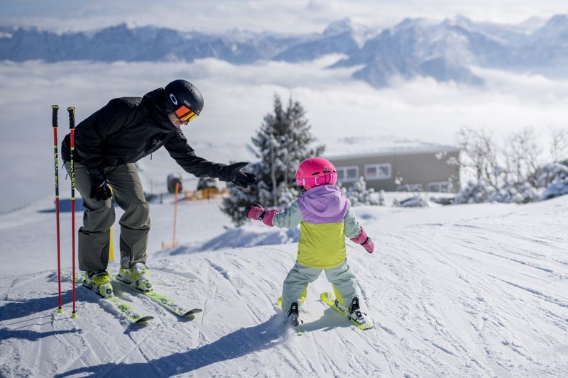 Kleinkind im Skianzug greift nach der Hand eines Erwachsenen auf der Piste, dahinter Berge und Nebelmeer.