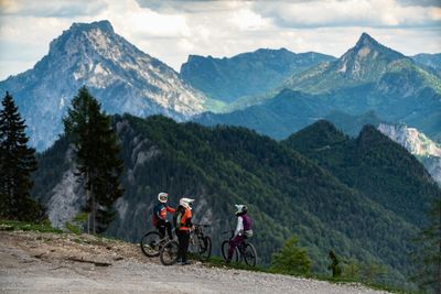 Drei Mountainbiker auf einem Bergweg mit atemberaubendem Blick auf bewaldete Berge und majestätische Gipfel unter bewölktem Himmel.