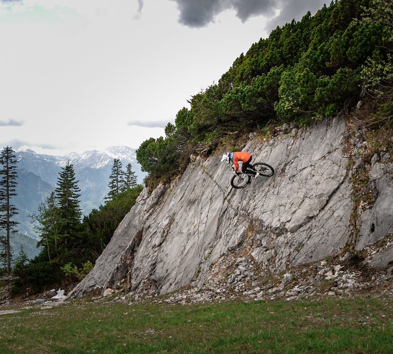 Mountainbiker in Orange überquert steile Felswand vor Bergpanorama.