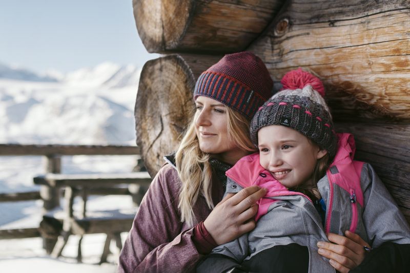 A mother cuddles with her daughter in warm winter jackets against a wooden hut, both smiling as they look into the distance.
