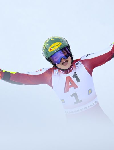 A female ski racer in a red racing suit stretches out her arms—ski poles included—in celebration.