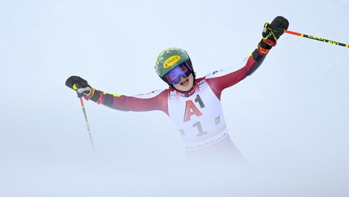 A female ski racer in a red racing suit raises her arms and ski poles in celebration.