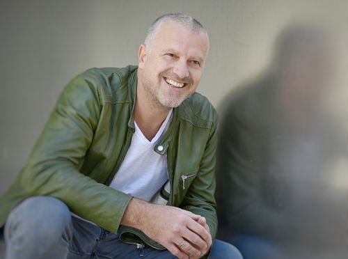 Erfolgsautor Bernhard Aichner sitzt lächelnd in grüner Jacke vor einer spiegelnden Fläche, in der sein Spiegelbild zu sehen ist.