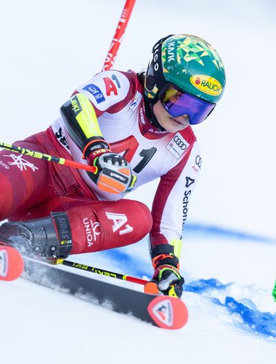Skirennläuferin in rotem Rennanzug bei tiefer Kurvenfahrt, Hand streift den Schnee, der aufspritzt. Grüner Helm und lila Brille.