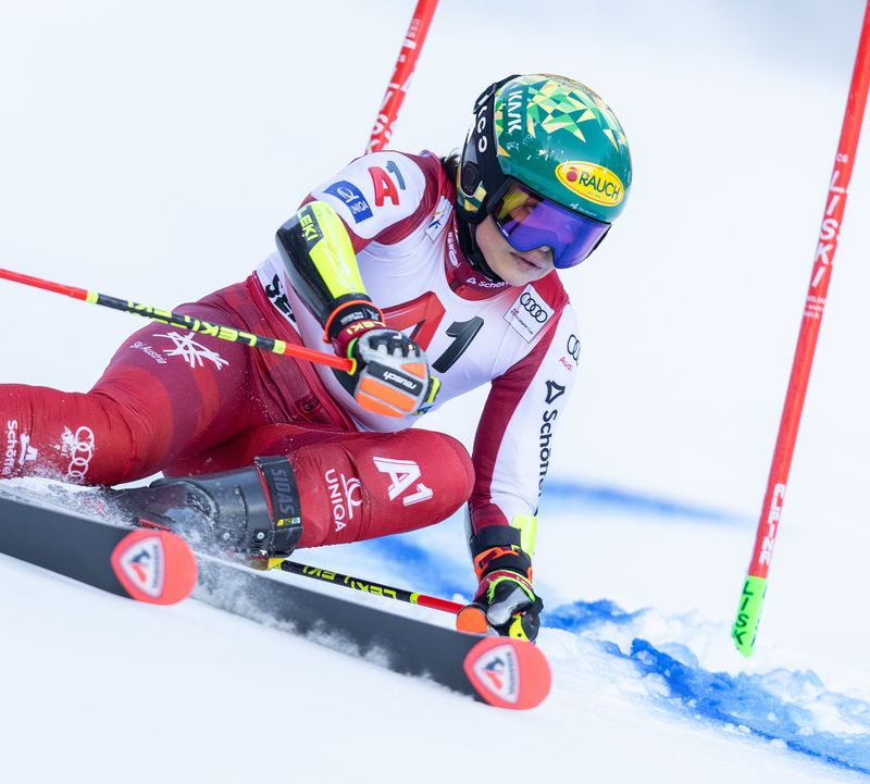 Skirennläuferin in rotem Rennanzug bei tiefer Kurvenfahrt, Hand streift den Schnee, der aufspritzt. Grüner Helm und lila Brille.
