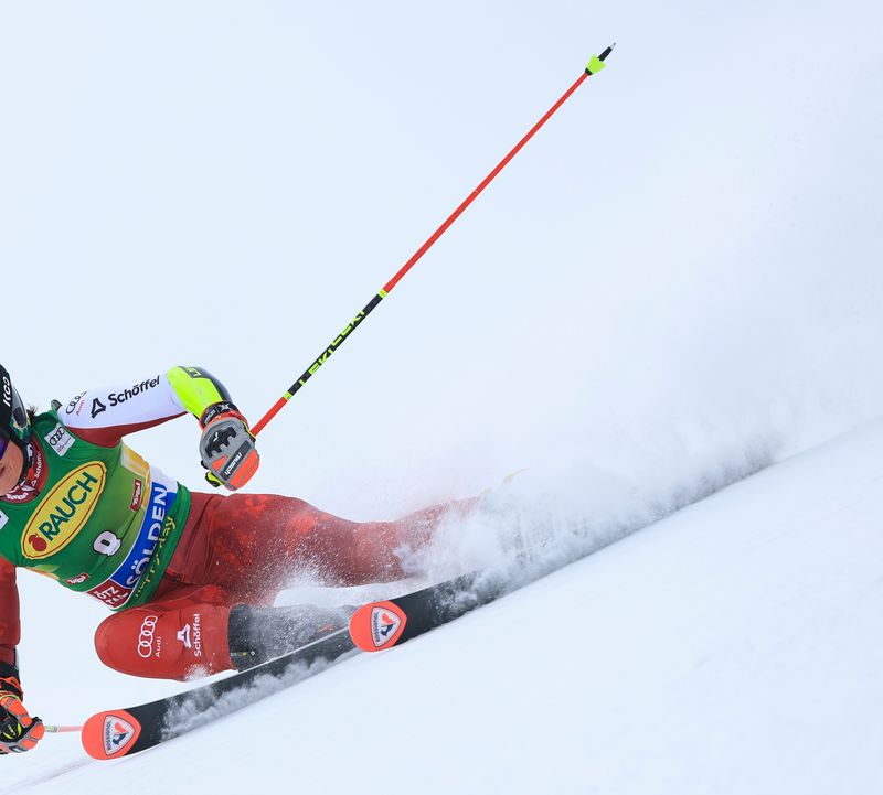 A female ski racer in a red racing suit skis across the snow-covered racecourse in a deep, banked turn, kicking up snow as she goes.