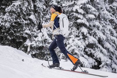 Person, die durch einen verschneiten Wald bergauf mit Skitourenski geht. Sie trägt eine Winterausrüstung trägt, darüber einen weißen Rucksack.