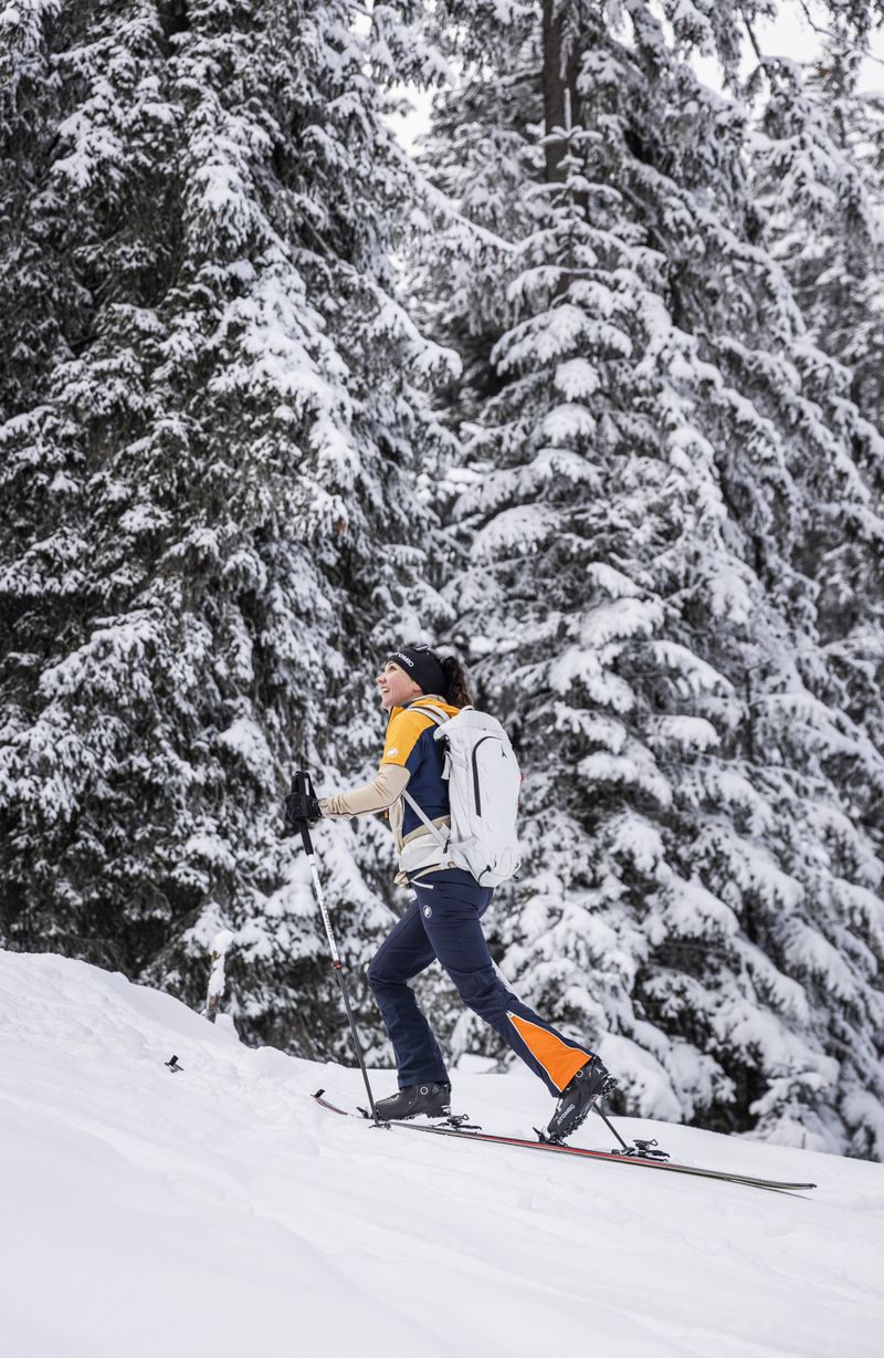 Person, die durch einen verschneiten Wald bergauf mit Skitourenski geht. Sie trägt eine Winterausrüstung trägt, darüber einen weißen Rucksack.