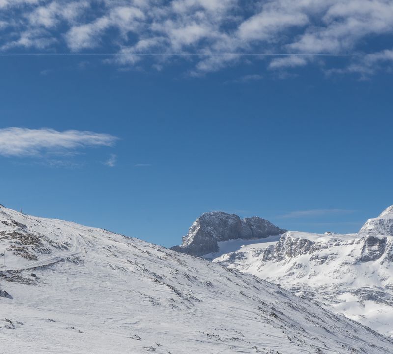 Snow-covered mountain landscape at Dachstein glacier with a clear blue sky and scattered clouds. A person stands on the left, silhouetted against the horizon.