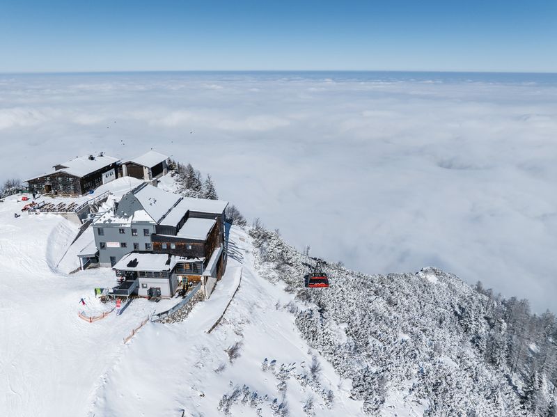 The mountain station on the Feuerkogel sits atop a snow-covered ridge, while a red gondola floats through the sea of clouds below.